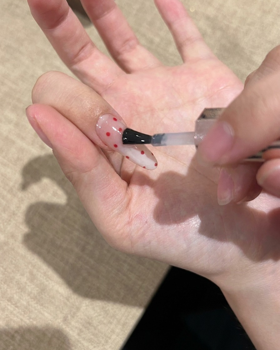 赤いドット柄のアートが施された爪に、クリアで艶のあるトップコートを塗布しているネイルブラシのクローズアップ。