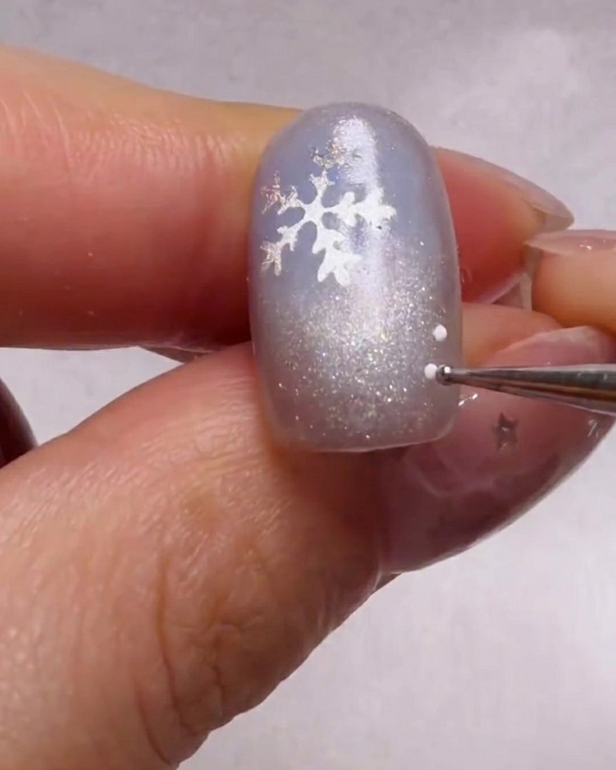 ドットペンを使って、きらめくブルーの爪にあるシルバーの雪の結晶ステッカーの周りに、白と水色の小さなドットを描いているクローズアップ画像。
