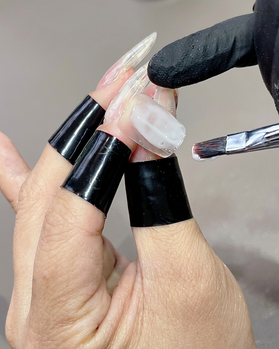 長いクリアネイルチップに、フラットブラシでミルキーホワイトのジェルベースを薬指に均一に塗布している様子。