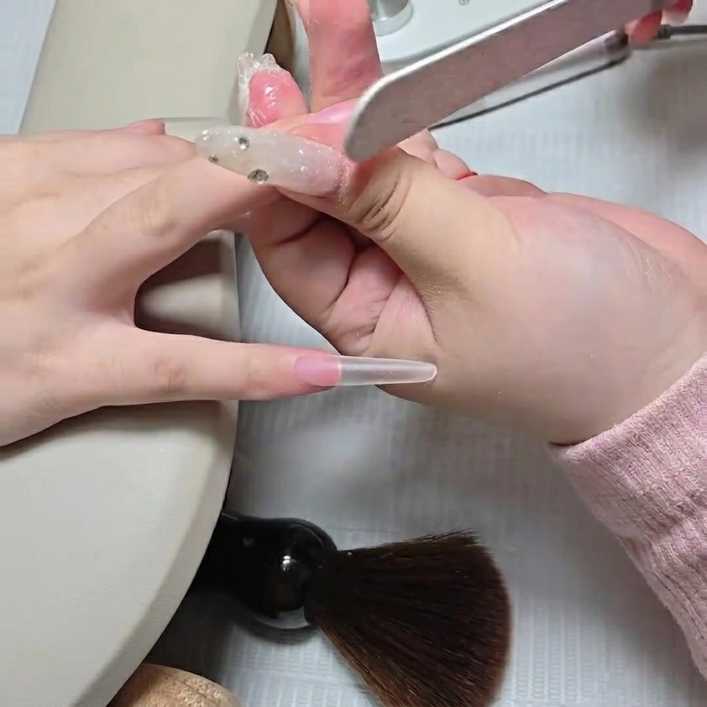 ネイリストがファイルを使って、自爪とクリアなスティレットネイルチップの境目の段差を削って滑らかにしている様子。