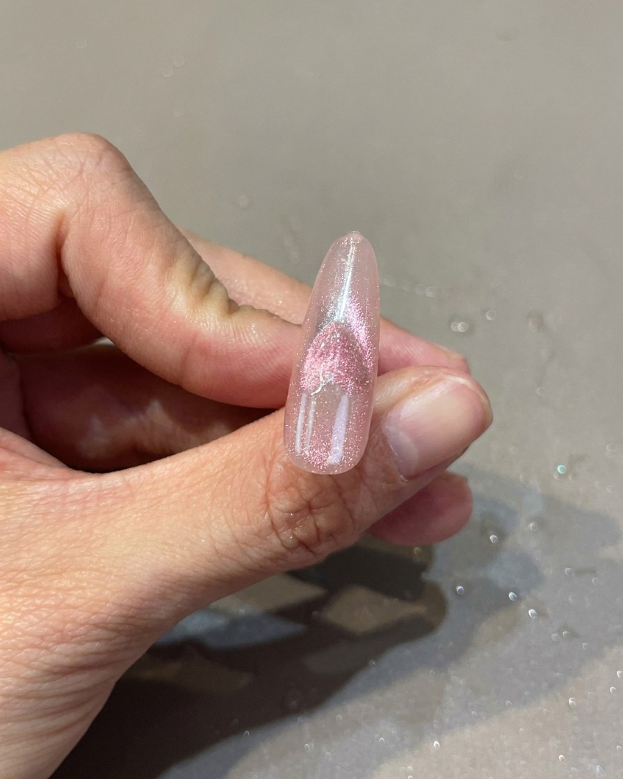 ピンクのネイルの上で、マグネットを使って磁石の粉を動かし、ハートの模様を浮かび上がらせている様子。