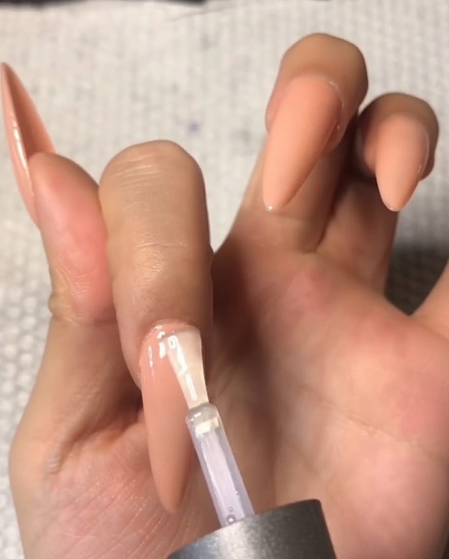 ヌードカラーのアーモンド型ネイルの甘皮周りに、テンパードトップコートを丁寧に塗布しているクローズアップ。