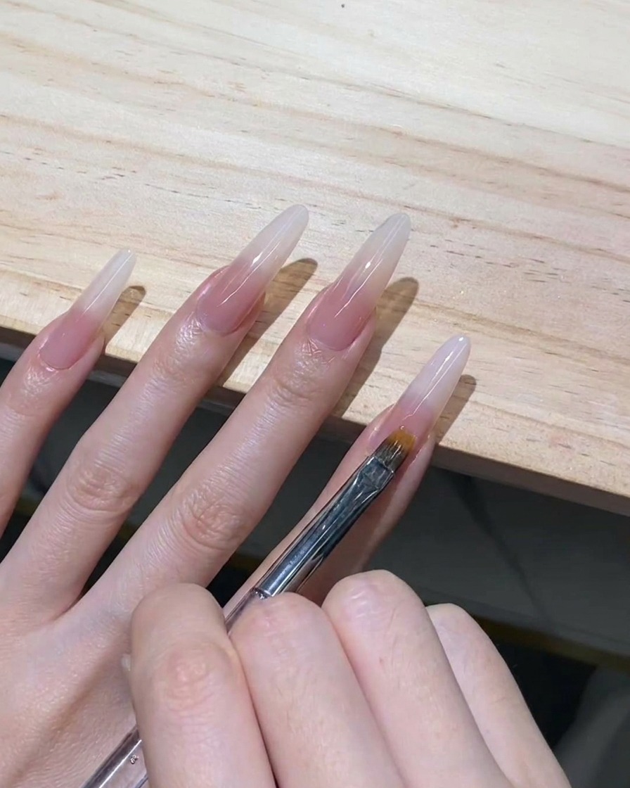 透明なスティレットネイルの先端に乗せたミルキーホワイトのジェルを、細いオンブレブラシで下に向かって優しく引いている様子。