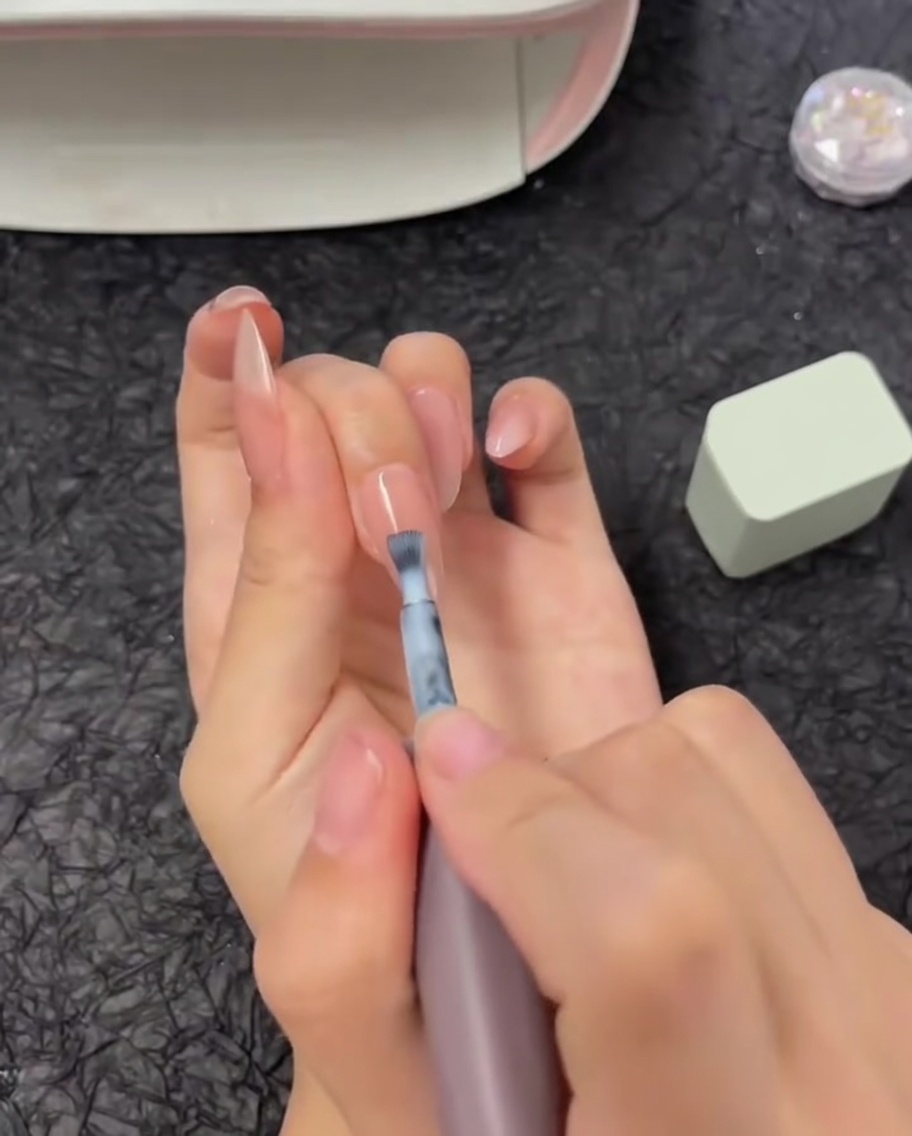 A brush applying a horizontal strip of white gel polish to the top half of a fingernail.