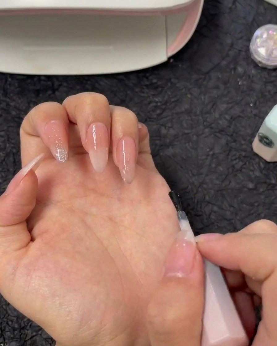 A brush applying clear reinforcement gel directly onto an index fingernail with a nude gradient base.