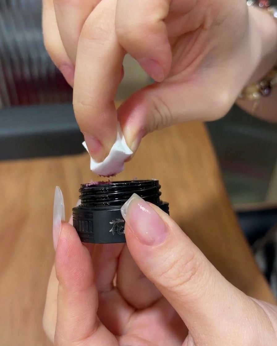 黒いコスメポットから、白いスポンジの角に少量の明るいピンクのジェルを取る手元のアップ。