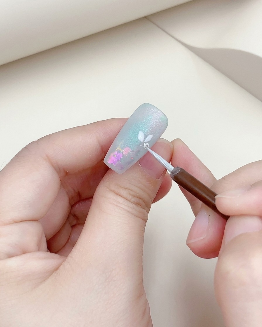 細いブラシで、ネイルに描いた白い花をシルバーで縁取っているクローズアップ写真。