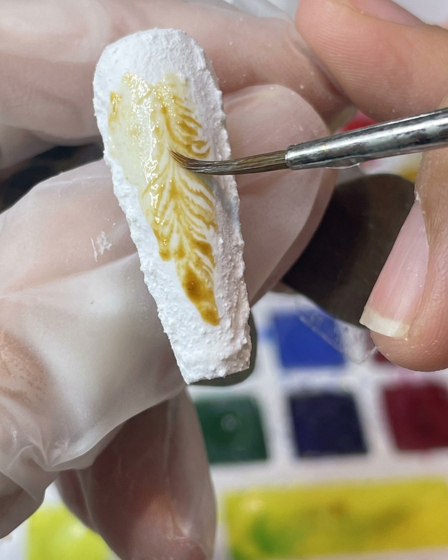 テクスチャーのある白いネイル表面に、黄色と茶色のピグメントを筆で塗布している様子。