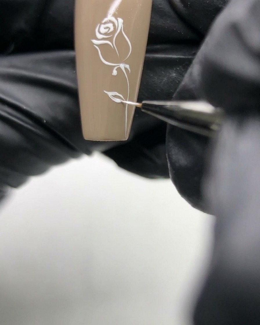 ニュートラルベースのネイルに描かれた薔薇の繊細なホワイトラインを、ネイリストが細かく整えているマクロ写真。