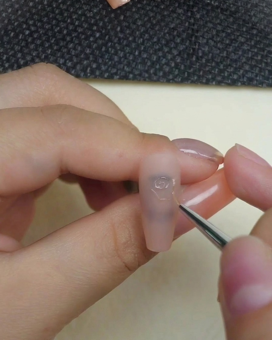 マットなヌードネイルの上に、細いライナーブラシを使ってバラの中心の渦巻きを3Dスカルプチュアジェルで描いている様子。