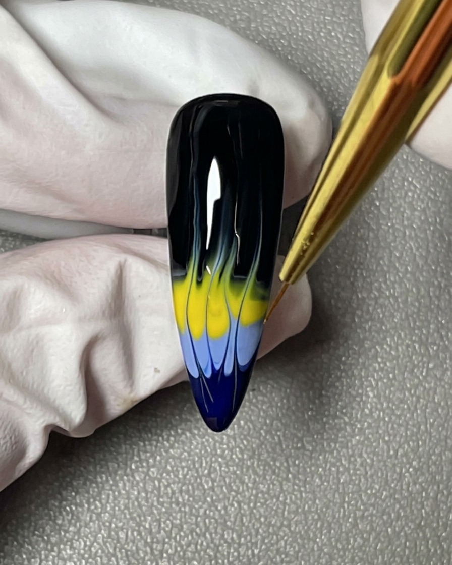 ライナーブラシで、黒い爪の上に描かれた青と黄色の炎の先を尖らせて整えている様子。