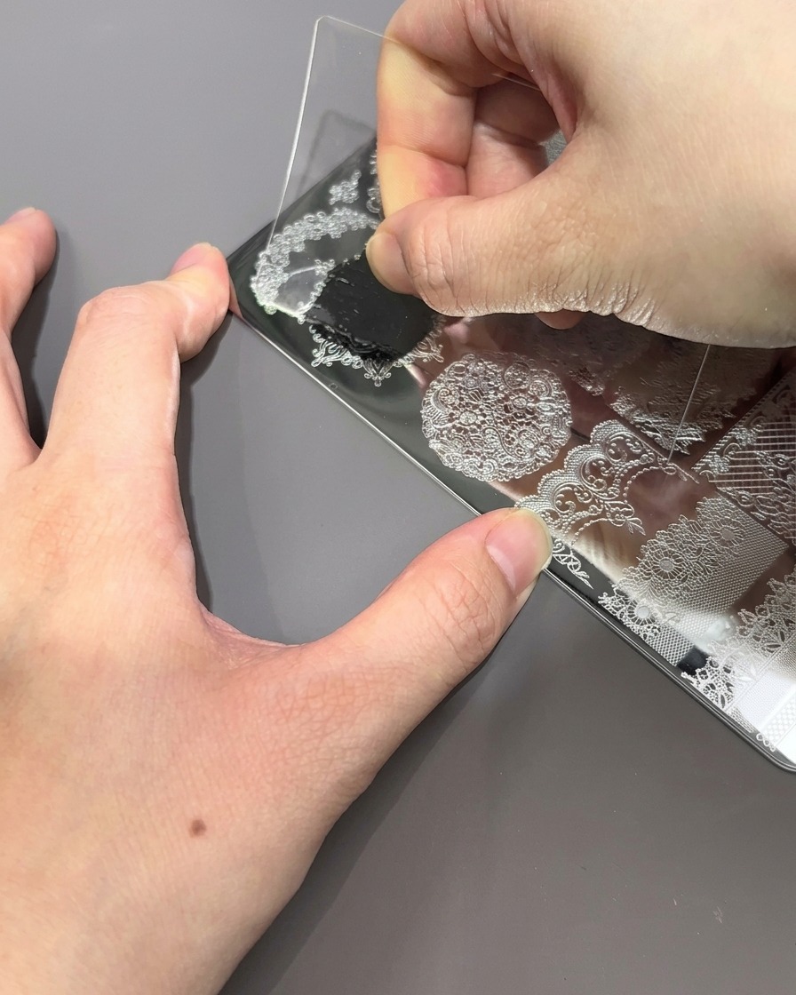透明なプラスチック製のスクレーパーカードをメタルプレートに押し当てて滑らせ、余分な黒のポリッシュを取り除いて模様を浮かび上がらせている様子。