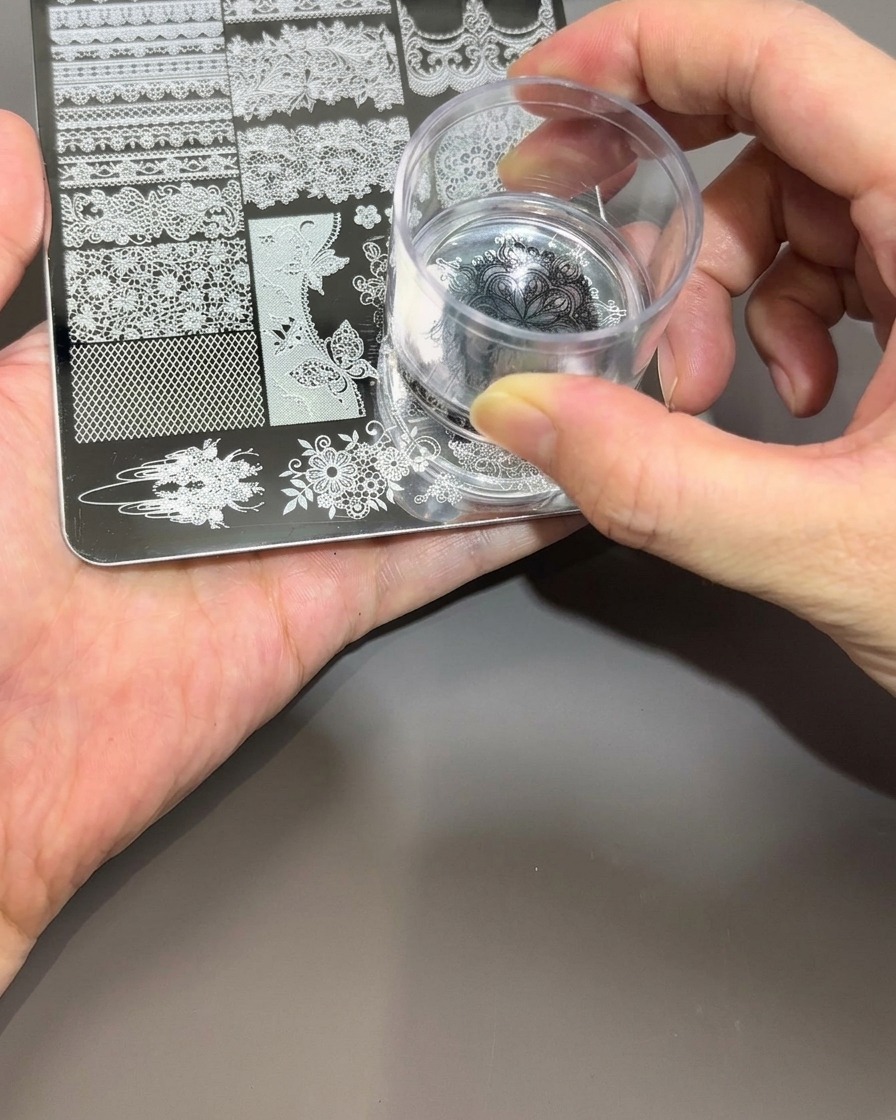 手に持った透明なシリコン製ネイルスタンパーを、メタルプレート上の黒いポリッシュ模様に優しく押し当てている様子。