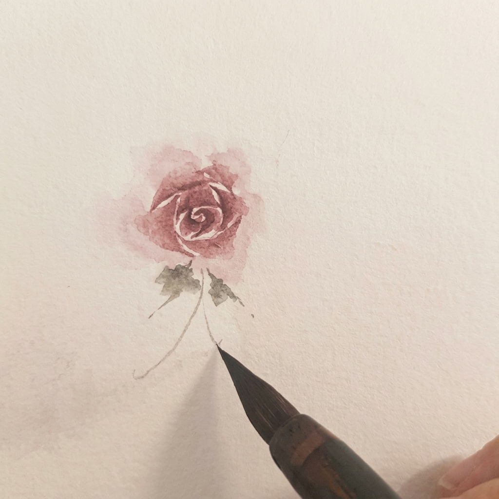 完成したバラの根元に、緑の水彩絵具で2枚の小さな葉が描かれている様子。