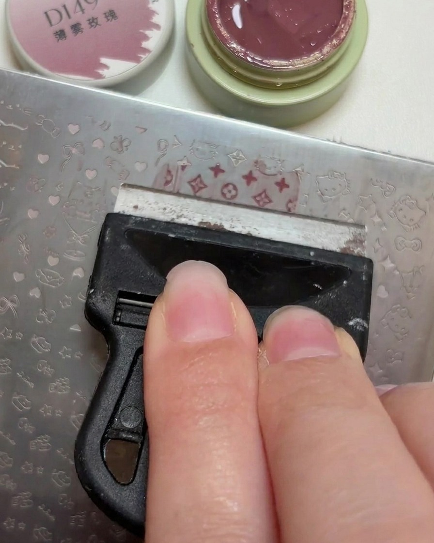 プラスチック製のスクレーパーを使って、金属製スタンピングプレートの彫り込まれた模様に沿ってダスティローズのジェルを均している様子。
