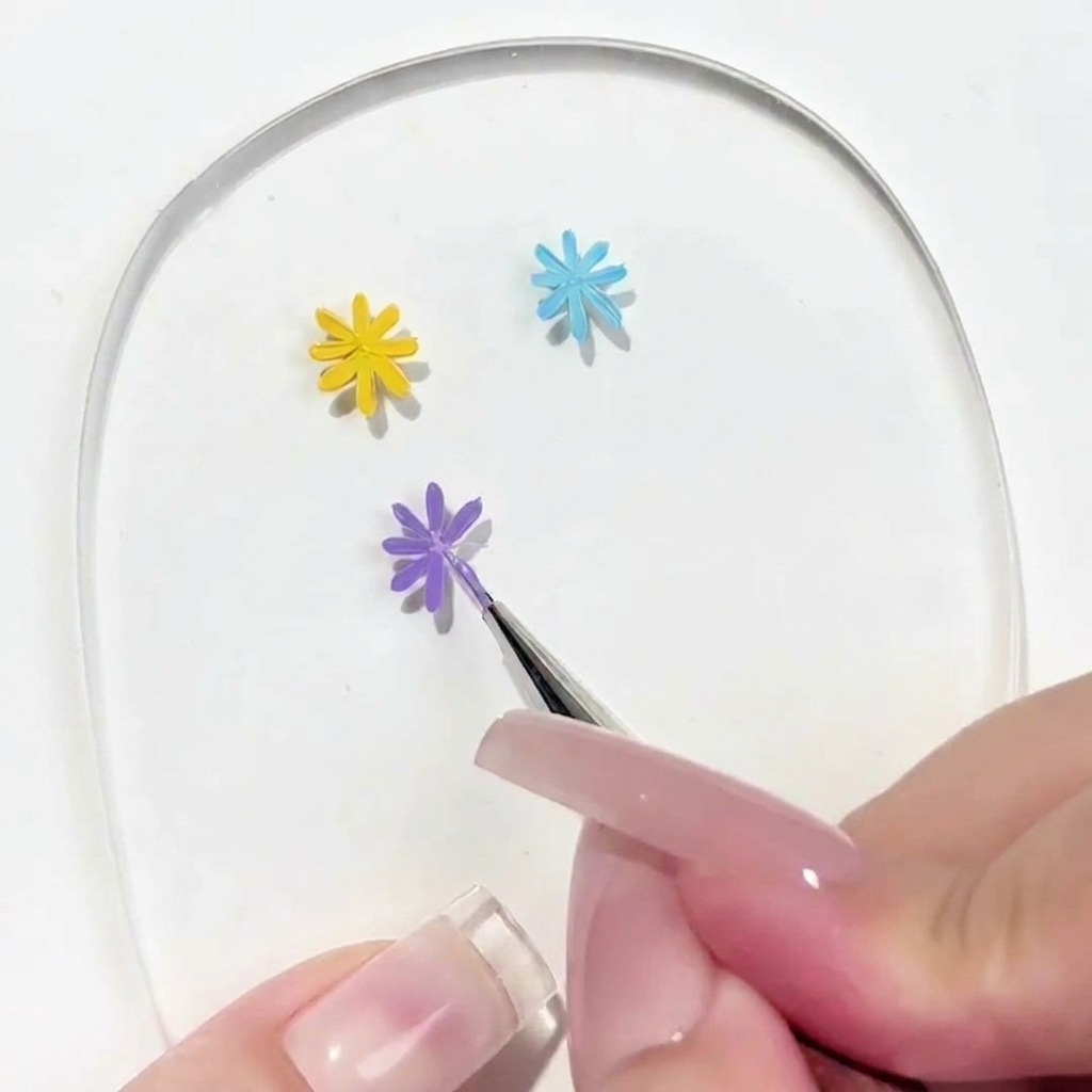 クリアなネイルパレット上で、描きかけのパープルデイジーに5枚目の花びらを足しているディテールブラシのクローズアップ画像。