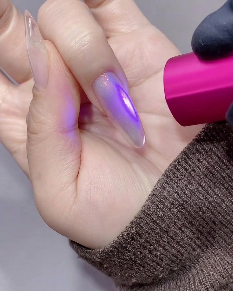 透け感のあるパープルがかった長いスティレットネイルを、小さな手持ち式のピンク色UV/LEDライトで硬化しているクローズアップ画像。
