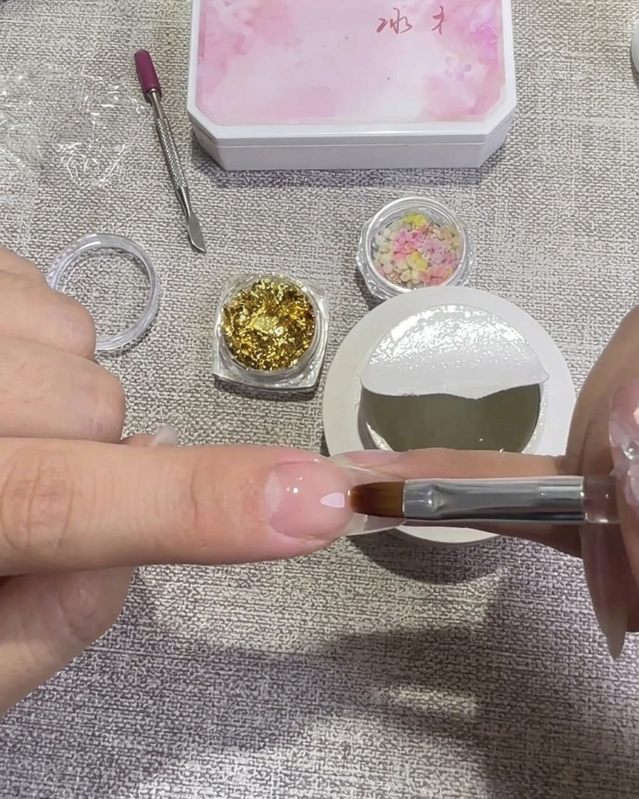 平筆を使い、クリアハードジェルを爪全体に塗っている様子。背景には金箔と押し花が入った小瓶が置かれています。