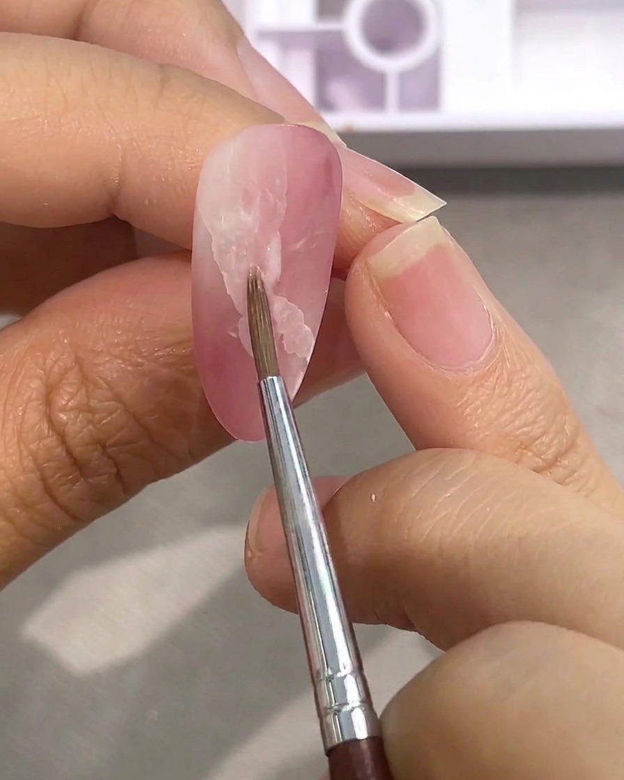 マットなピンクネイルに乗せた白い水彩絵の具の輪郭を、水を使って細筆でぼかす様子
