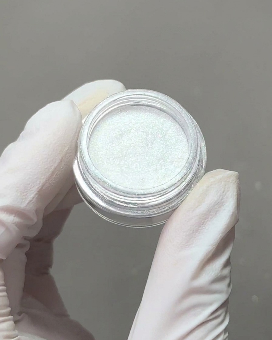 手袋を着用した手で、繊細なオーロラクロームパウダーの容器を持ち、アイシャドウチップで少量のパウダーをすくい取っている様子。