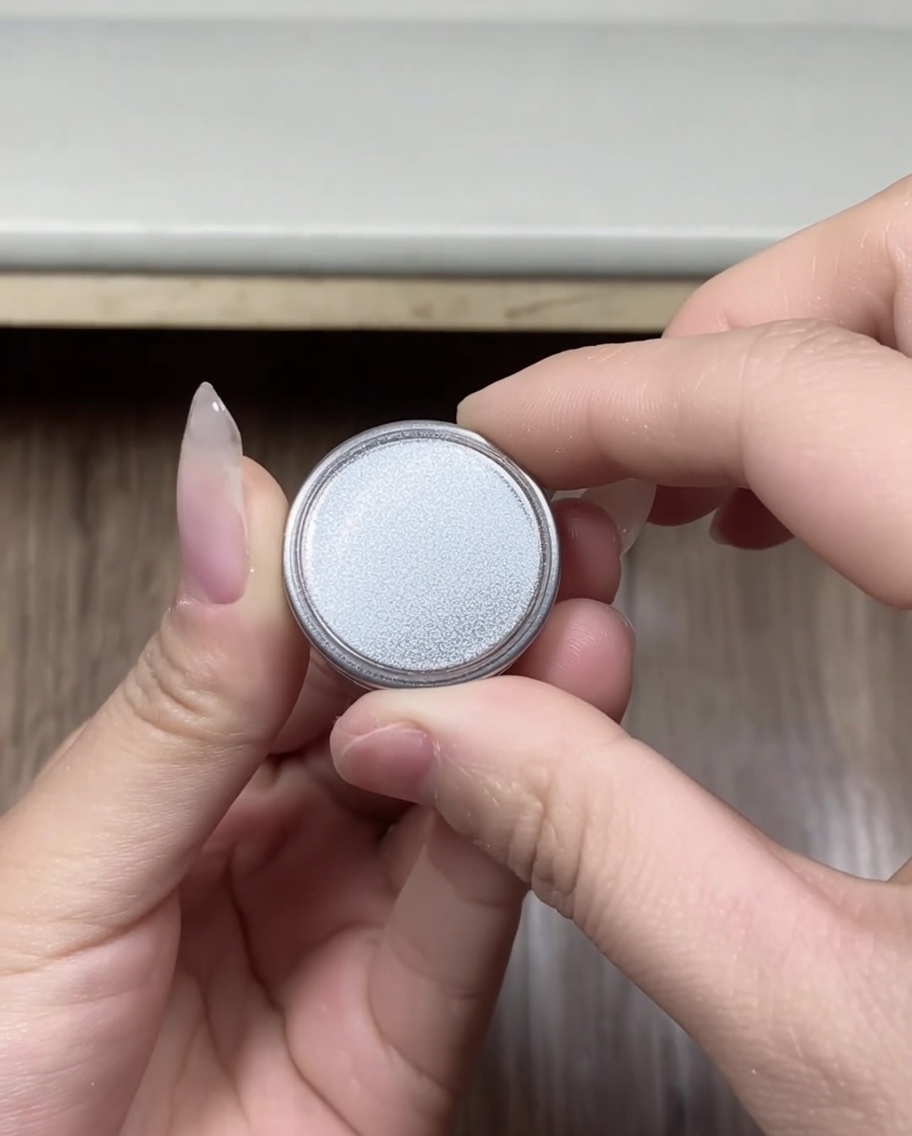 蓋を開けたばかりの、シルバーのクロムパウダーが入った小さな丸い容器を持っている手のクローズアップ。