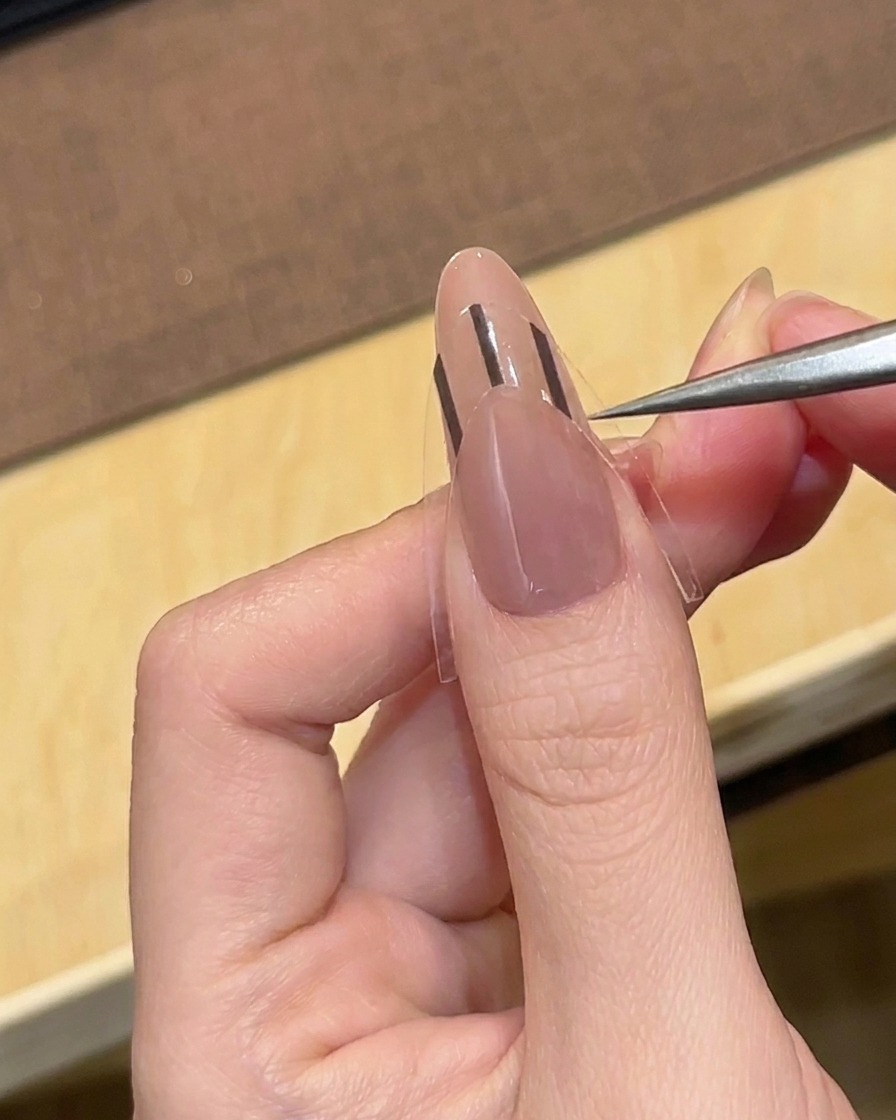 ピンセットを使って、ヌードカラーのアーモンドネイルに黒いフレンチガイドシールを貼っている様子。