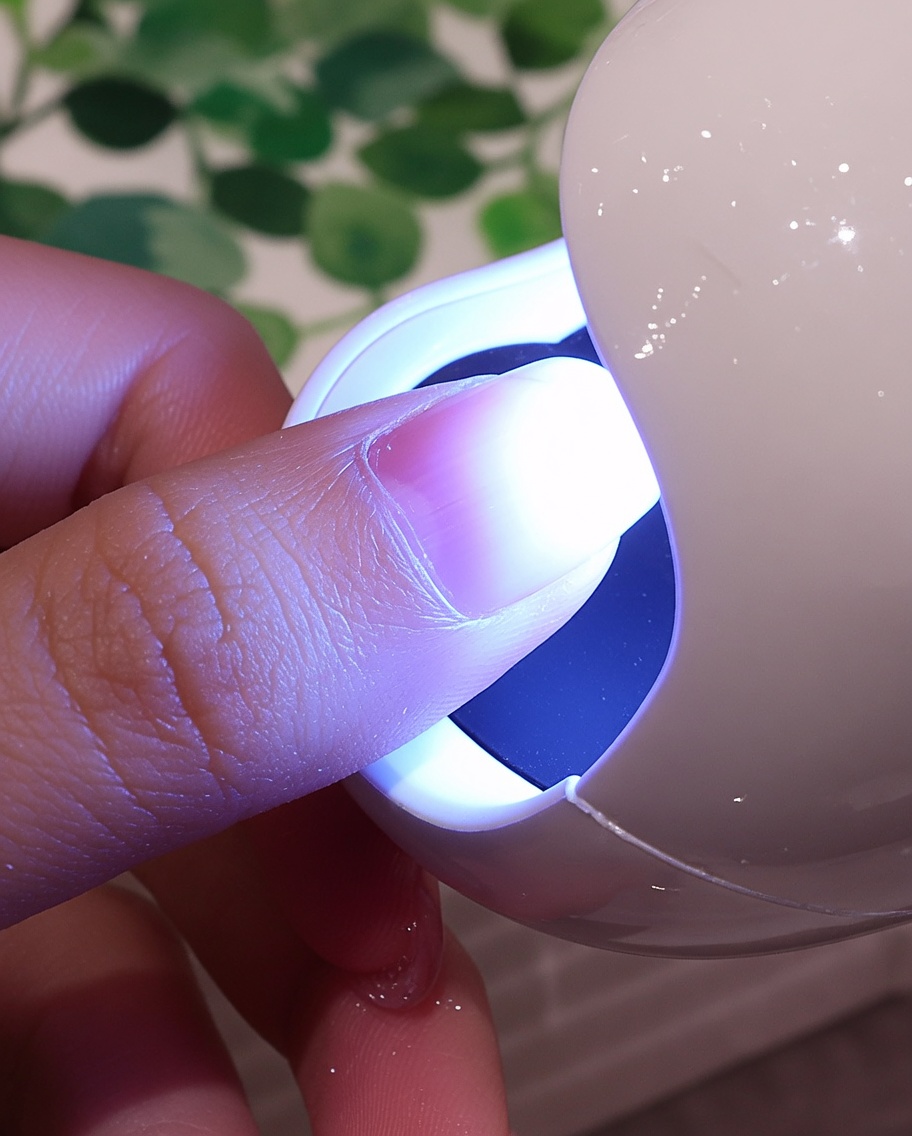 白いベルベット素材で覆われた爪を、小型UV/LEDネイルライトの紫色の光の下に配置している様子。