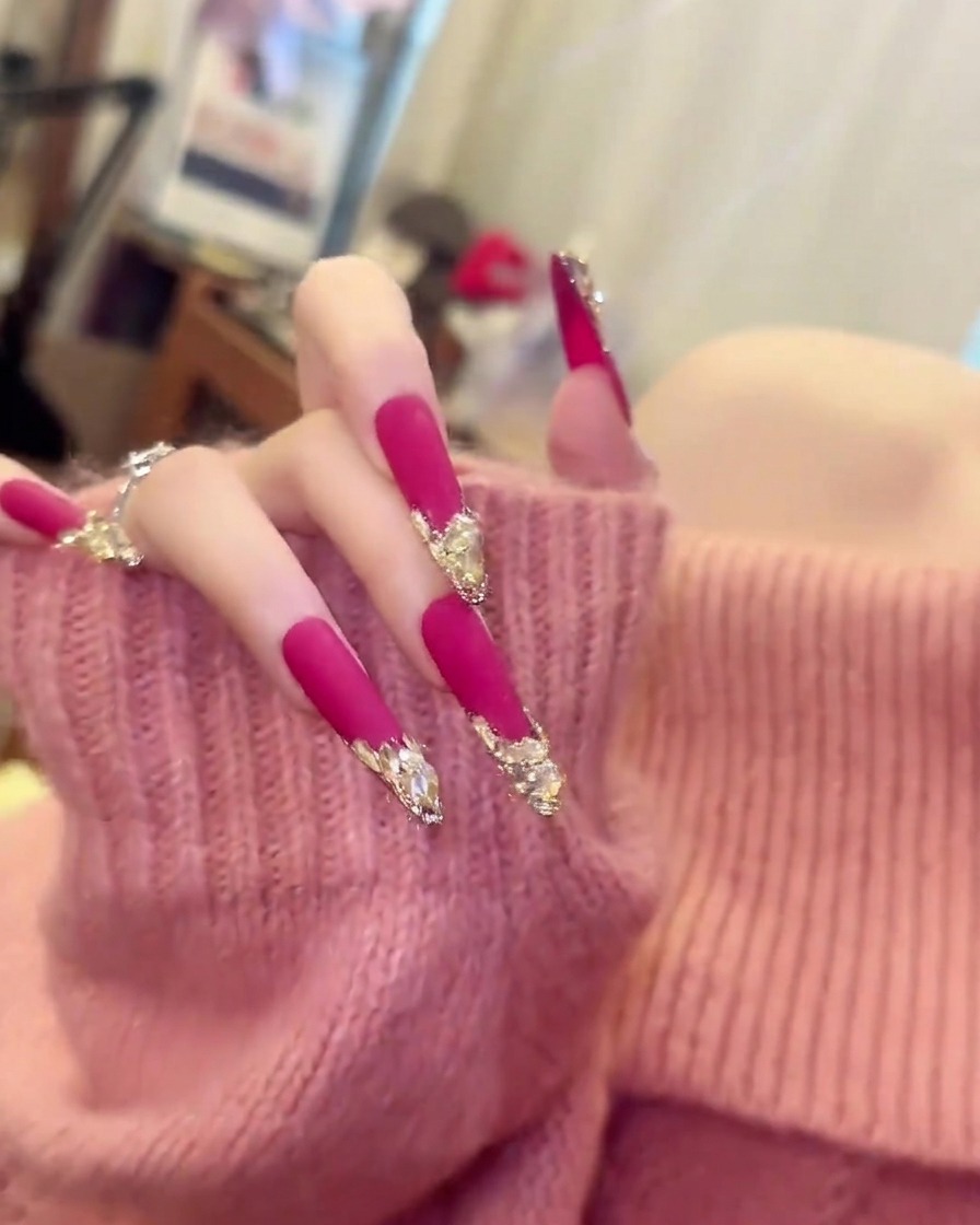 柔らかいピンクのセーターを着た手がゆっくりと動き、長いマットピンクのアーモンドネイルの長さとカーブを見せている様子。