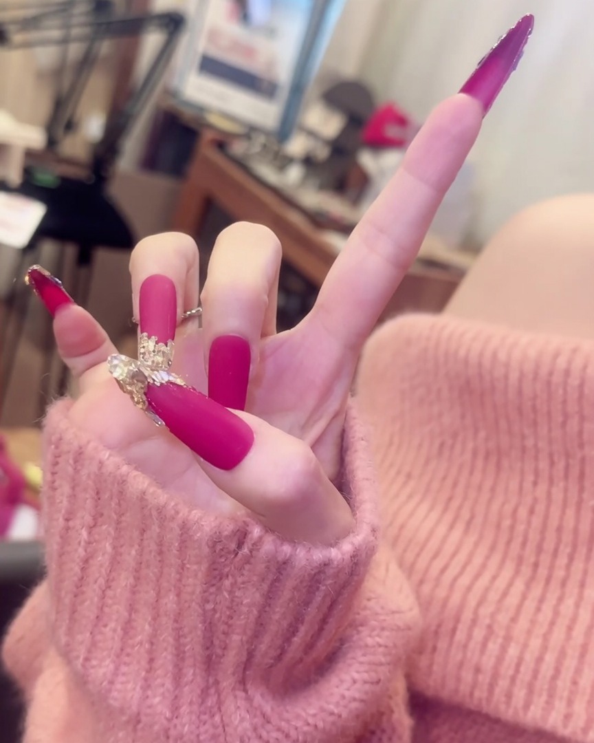 指を広げた手。エレガントなシルバーのリングアクセサリーとともに、クリスタルがアクセントになった長いマットピンクのネイルを披露している様子。