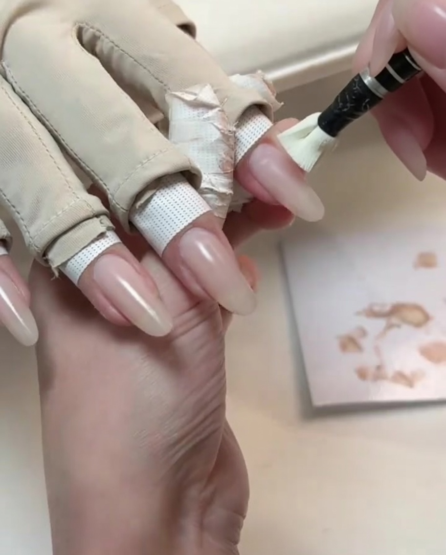 ロングのナチュラルな形の爪に、ライトヌードのジェルポリッシュを小さなアプリケーションブラシで塗布している様子。