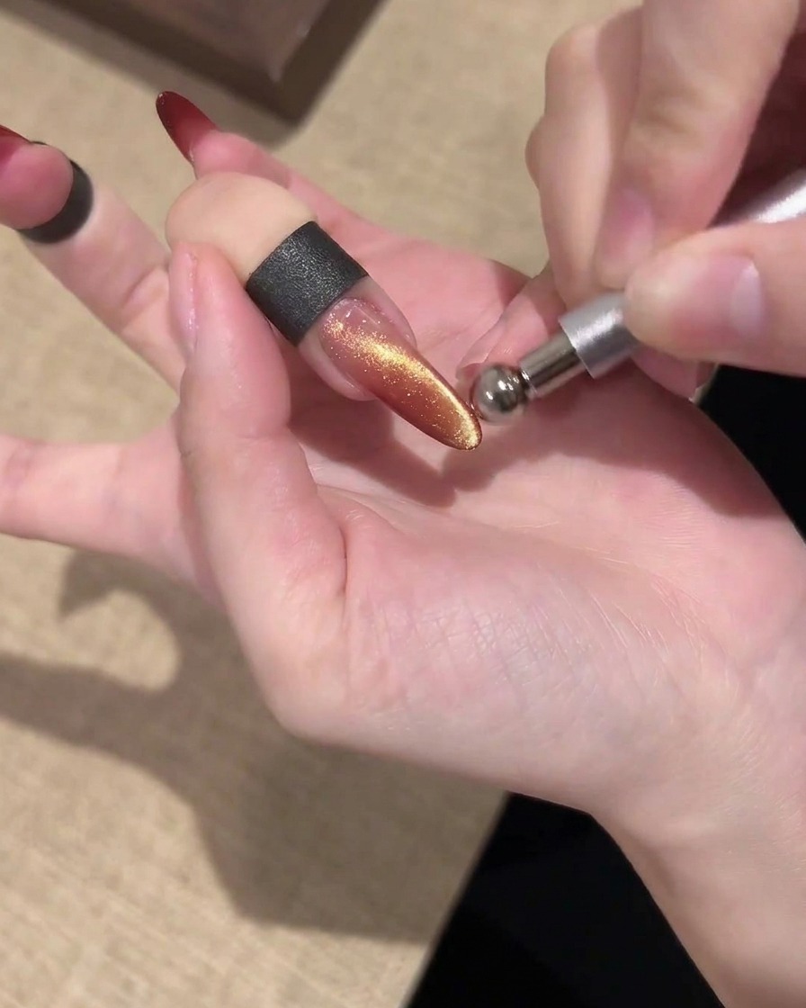 光を形作るために、きらめくゴールドとレッドのキャッツアイネイルの上方に平行に保持されたバーマグネット。
