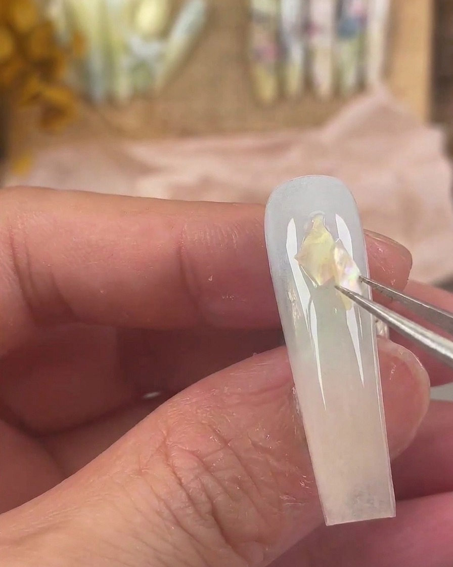 先細のピンセットで、濡れたジェルの表面にイリデッセント（偏光）なシェルの破片を配置している様子。