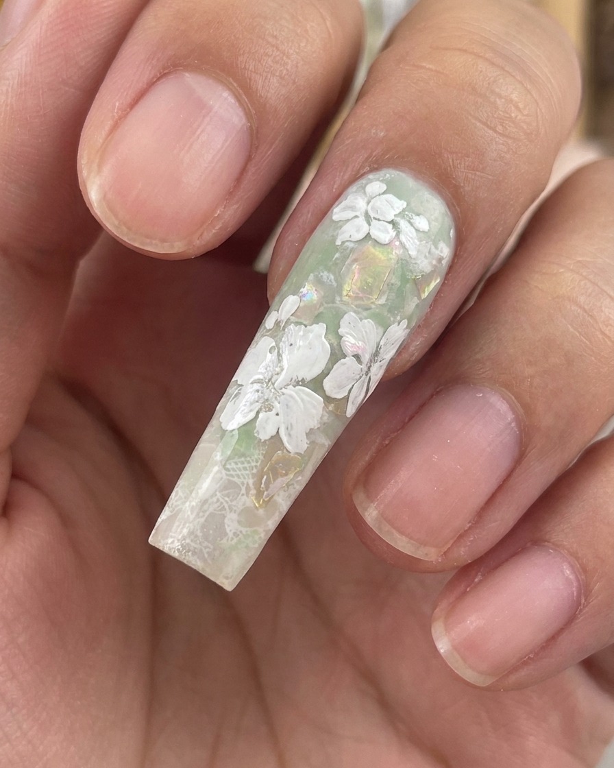 グリーンとシェルフレークの背景の上に、完成した白いジェルの花と小さな葉が見えるロングスカルプネイルのクローズアップ。