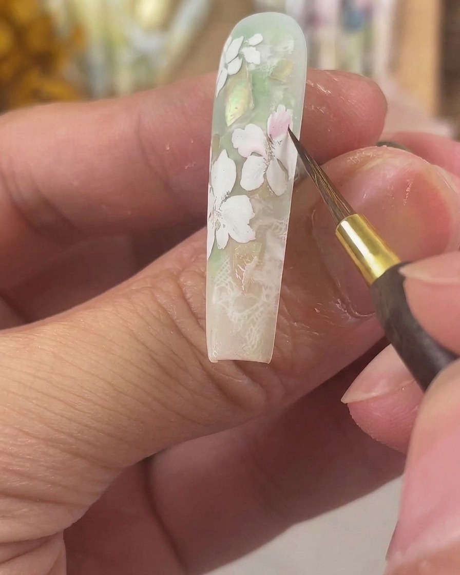 極細のディテールブラシで、ロングスカルプネイル上のマットな白いジェルの花びらにシアーなピンクの水彩顔料を塗布している様子。