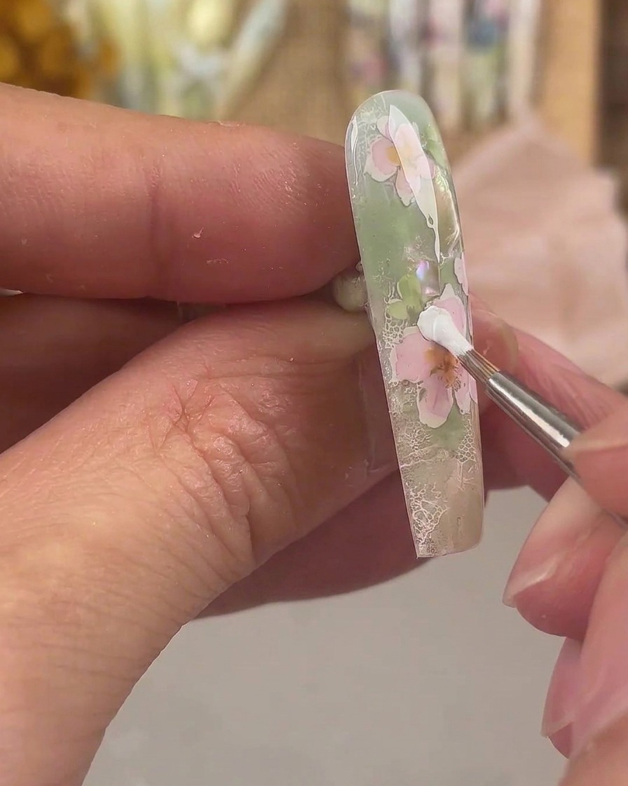 極細のライナーブラシで、ピンクのフラワーネイルアートのデザイン上に白いペイントジェルを使って余分な層の花びらを描いている様子。