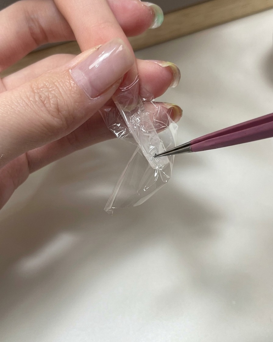 ピンセットを使用して、透明なネイルエクステンションの裏側に透明なプラスチックラップの小片を押し込み、しわのある質感を創り出しているクローズアップ。
