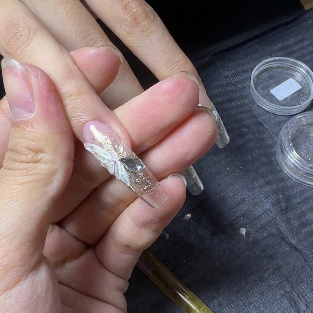 3Dの白い蝶の羽と、その脇に配置されて蝶の体を形作る2つのフラットバックラインストーンが特徴のクリアネイルエクステンションのクローズアップcite: 1。