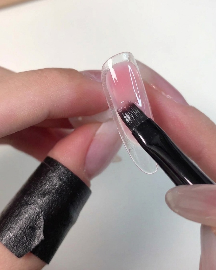クリアなネイルチップを持ち、フラットブラシでピンクの未硬化ジェルの端を優しくぼかしている手元。