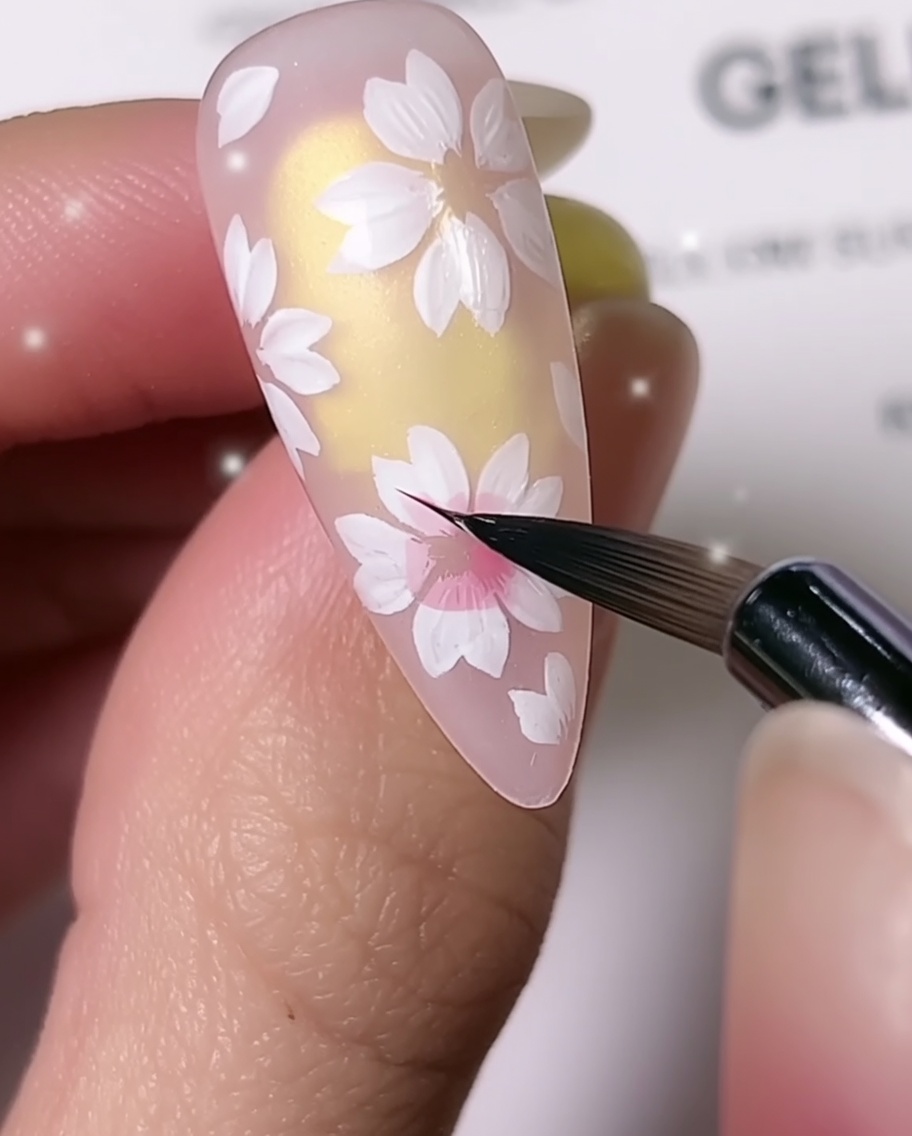 アーモンド型のネイル上で、細かいディテールブラシを使用して白い桜の花びらの中心にピンクの陰影を加えているクローズアップ画像。