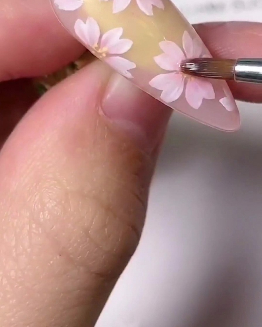 ネイル上のペイントされたピンクの桜の中心に、細かいディテールブラシ小さなゴールドの中心アクセントを適用しているクローズアップ画像。