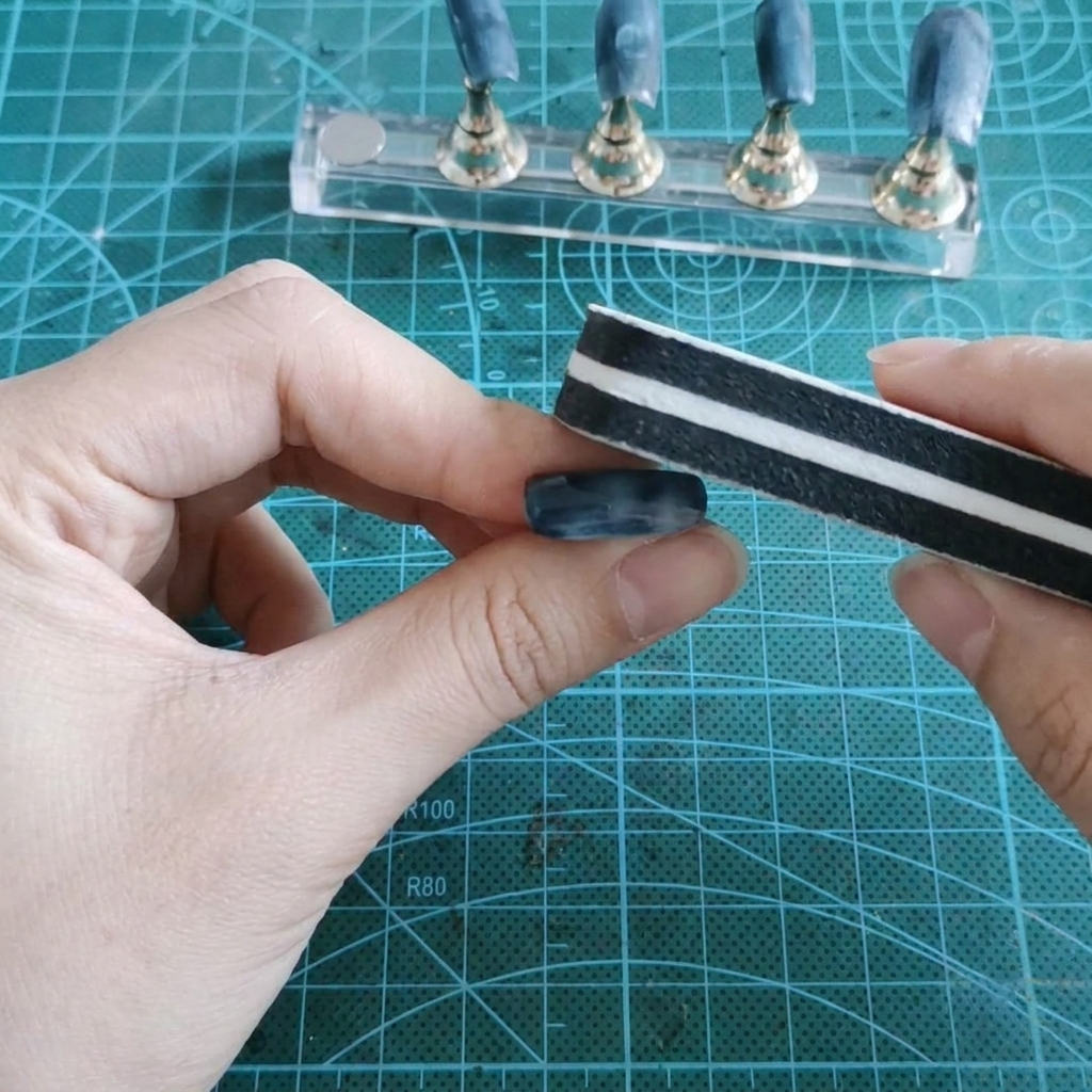 指の間に挟んだ偽のネイルの光沢のあるジェル表面を、ブロック型のスポンジバッファーでサンディングしている様子。