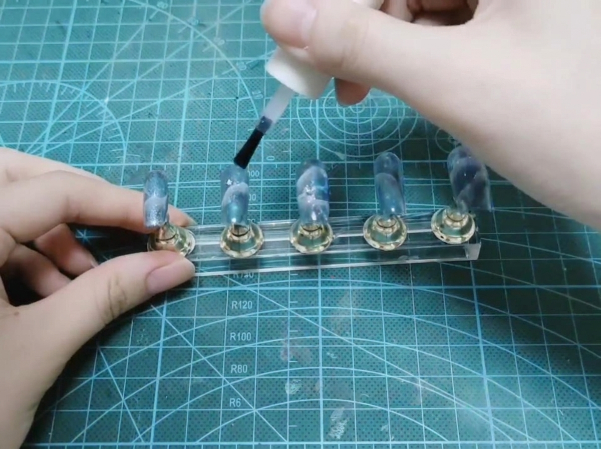 白いボトルのブラシを使って、ディスプレイスタンド上の完成したネイルセットの上に艶やかなトップコートを塗布している手。