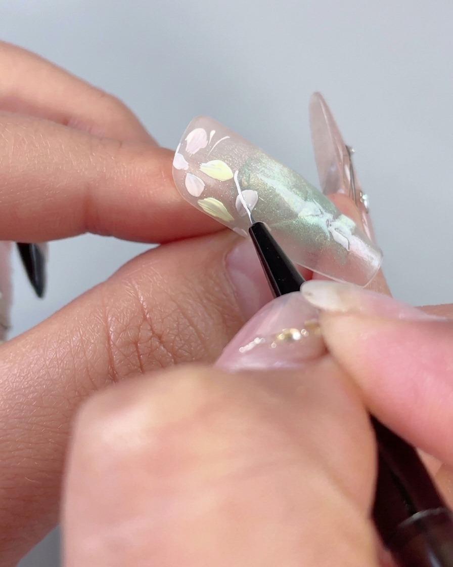 半透明のグリーンキャッツアイネイル上のパステルの花の隣に、細いライナーブラシが繊細な白い湾曲した線を描いている様子。