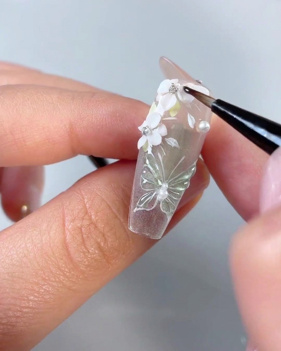 クリアネイルチップ上の白い3Dフラワーの花びらとラインストーンのエッジの周りに、細かいディテールネイルブラシがクリアジェルを適用している様子。