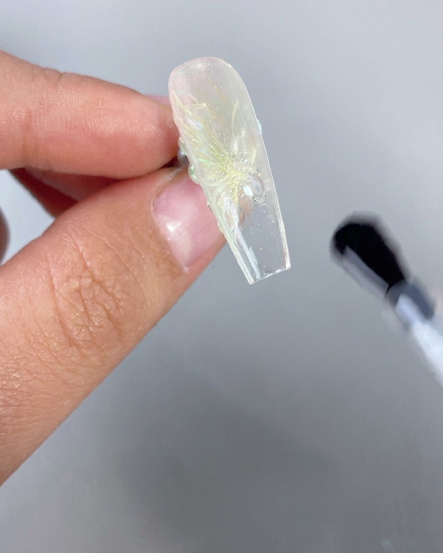 3Dフローラルデザインを持つアイス透明感のネイルを上下逆さに保持している手のクローズアップ。一方、ブラシがフリーエッジの裏側にクリアトップコートを適用している。