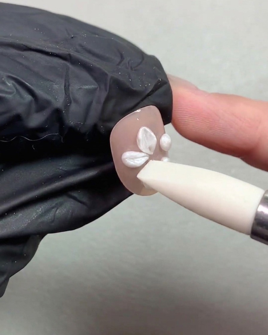 シリコンペンツールが、ヌードカラーに塗られた爪の上で白いパールのジェルボールを押さえて花びらに形作っている様子。