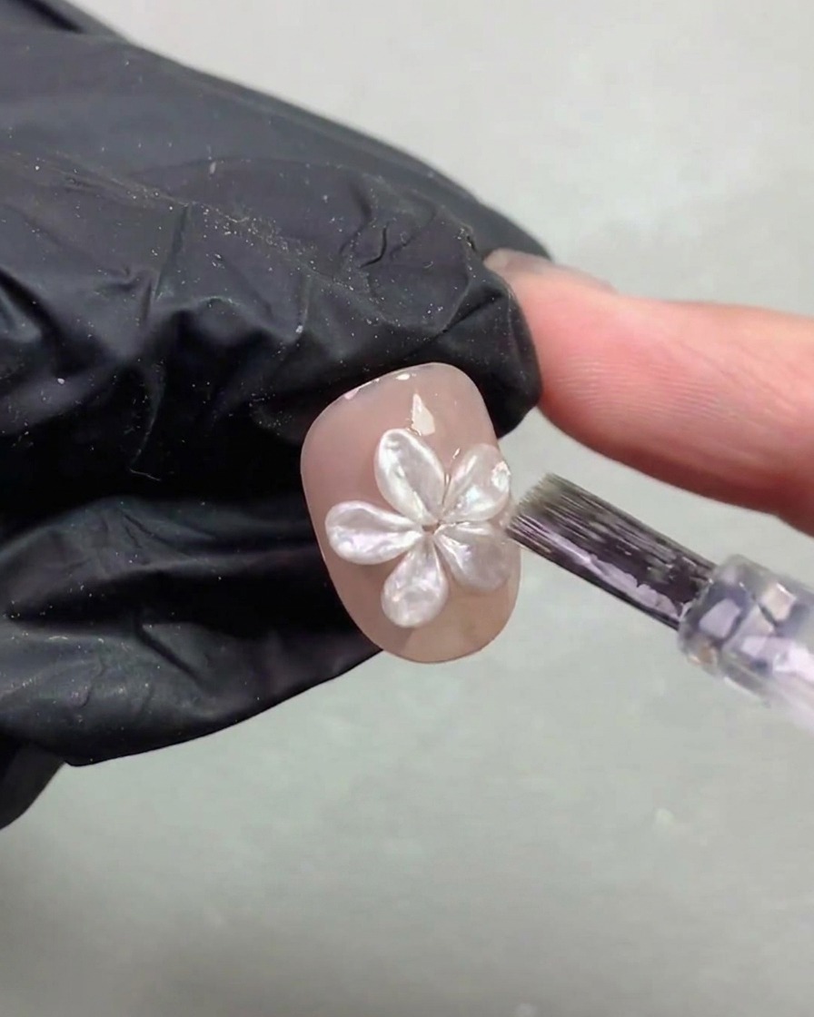 ネイルブラシが、硬化した3Dパールフラワーの周りに薄いトップコートを慎重に塗っている様子。