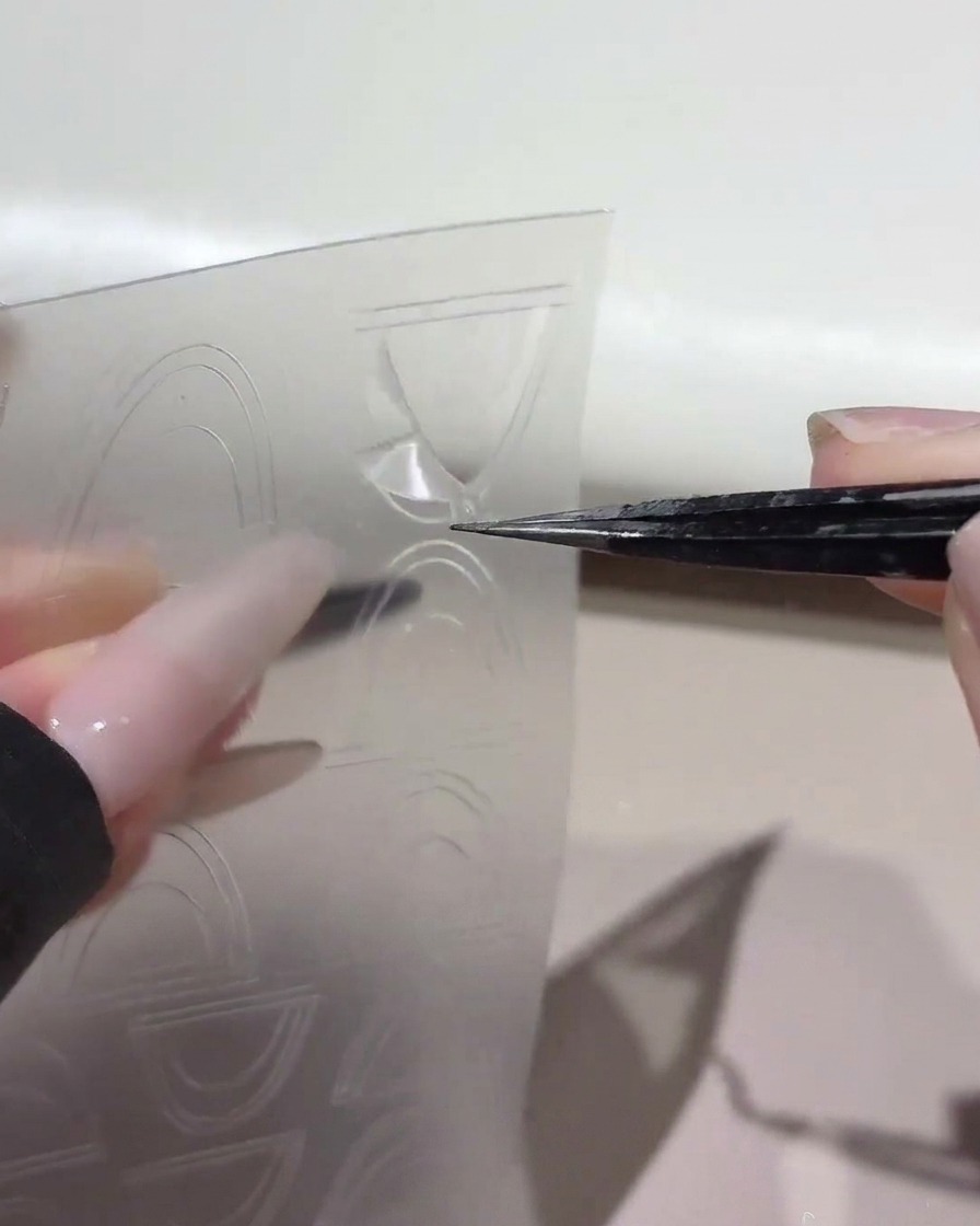 ピンセットを使って、透明なプラスチックシートからカーブしたフレンチガイドステッカーを持ち上げている様子。