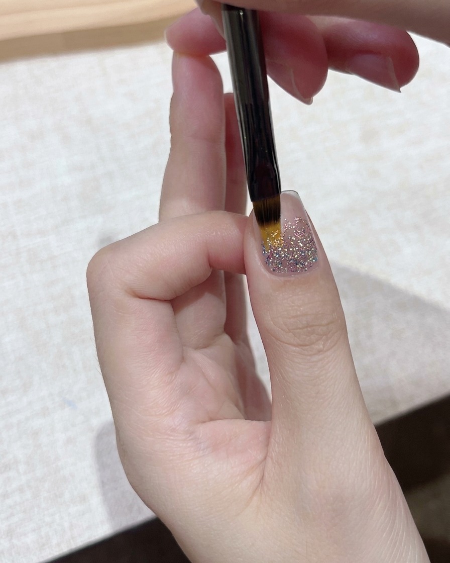 きらめくグリッターネイルのベースの上に、細筆を使ってクリアジェルの層を丁寧に塗布しているクローズアップ。
