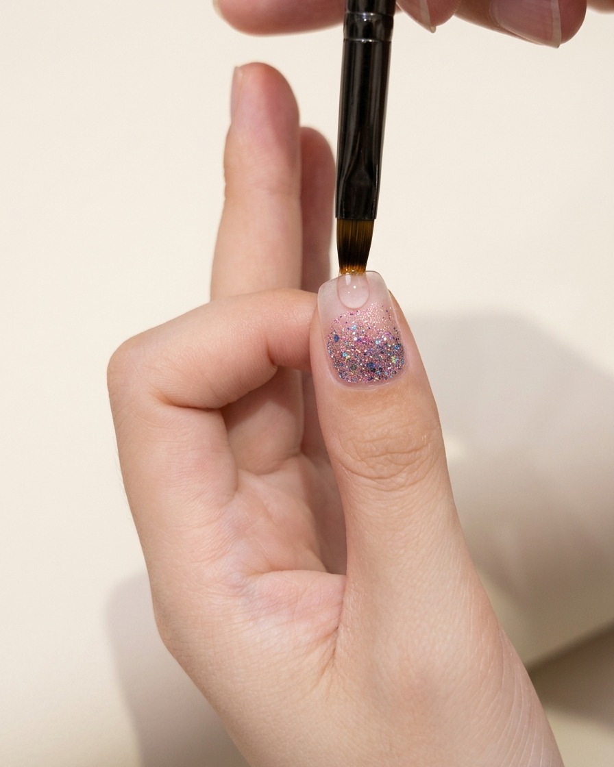 温かい光の下でグリッターのグラデーション層を確認している親指の爪のクローズアップ。